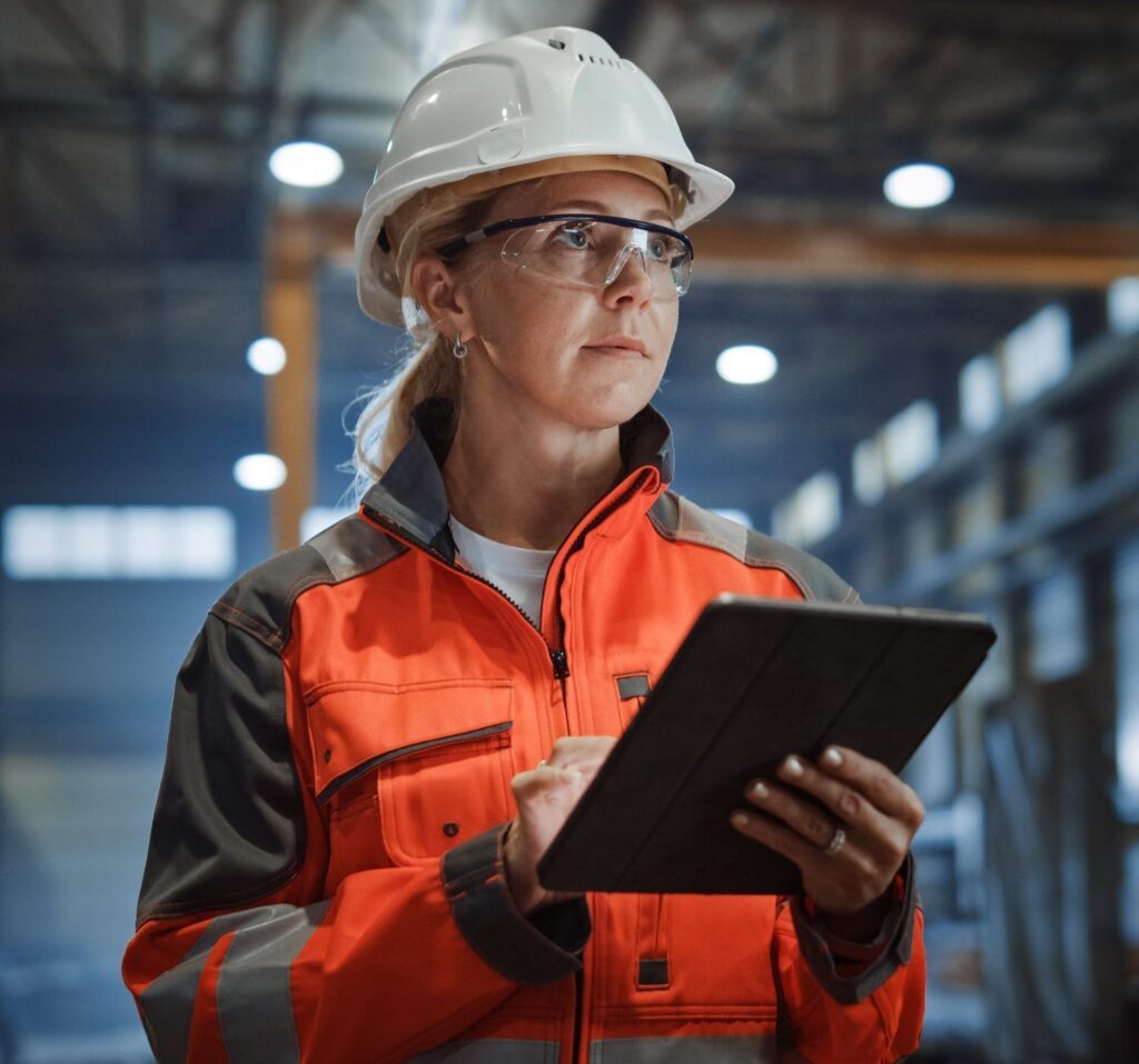 A woman wearing a high-visibility jacket, hard hat and saftey glasses stands in a factory enviroinment holding a computer tablet