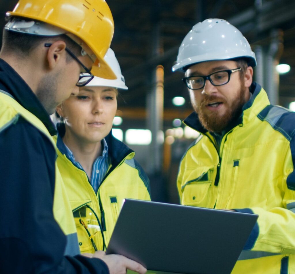 Two men and a woman in high visibility jackets and hard hats stand looking at a checklist in a factory environment
