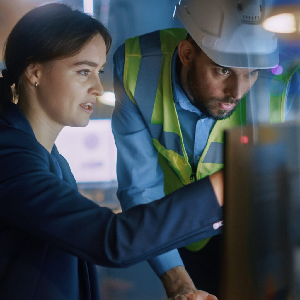 A woman in suit points at a computer screen as she explains something to a man in a high visibility jacket and hard hat who is standing next to her
