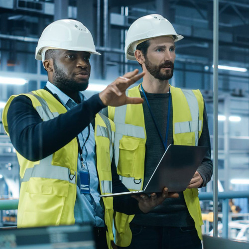 Two men in high-visibility jackets and hard hats stand in an industrial environment holding a laptop and discussing the plant machinery