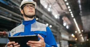 A man in a hard hat and blue overalls holding a tablet and looking round a factory