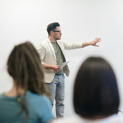 A man points at a whiteboard while teaching to a room of students