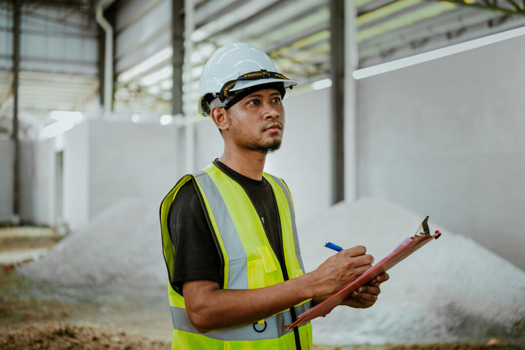 A man wearing a high-visibility jacket and hard hat stands in a storage facility holding a clipboard