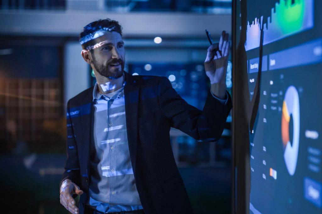 A man stands in the light of a projection indicating items on a projection screen