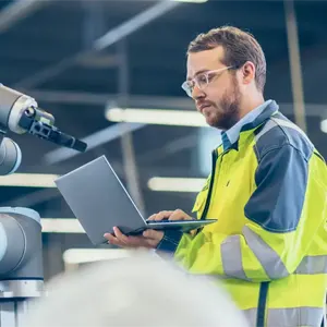 A man wearing a hi-viz jacket holds a laptop in one hand and types on it with the other, standing in an industrial environment