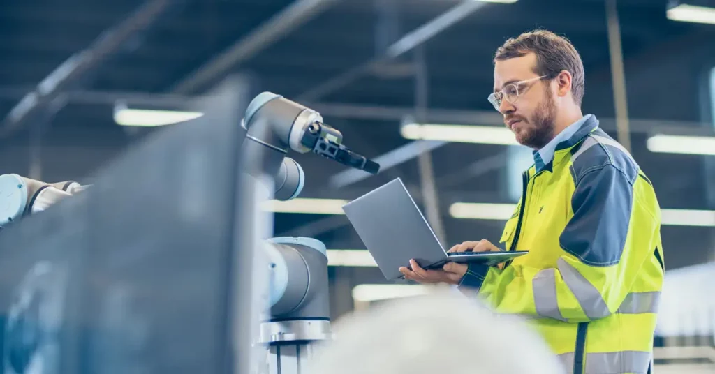 A man wearing a hi-viz jacket holds a laptop in one hand and types on it with the other, standing in an industrial environment