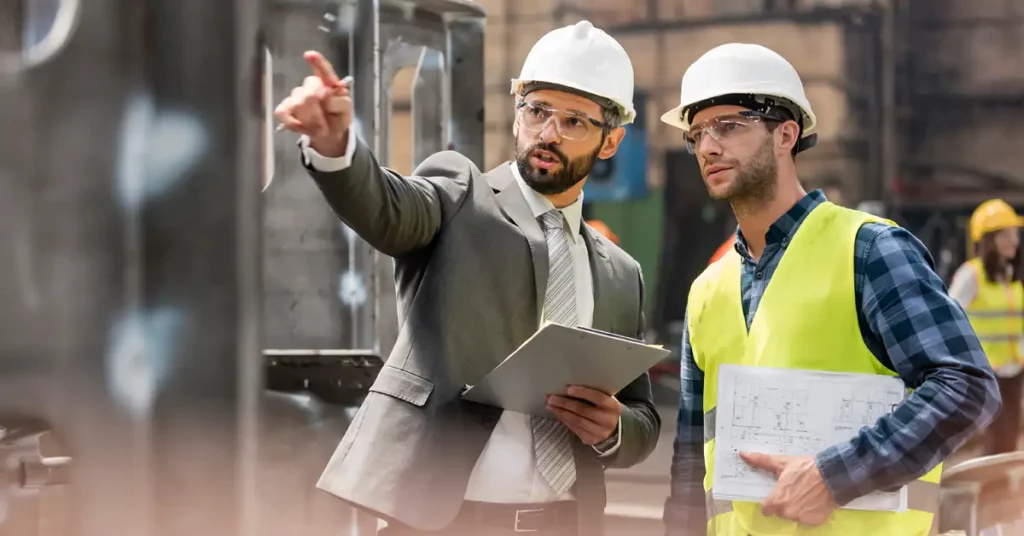 An external setting where a man wearing a suit and a hard hat is pointing something out to another man wearing a hard hat and a hi-viz jacket