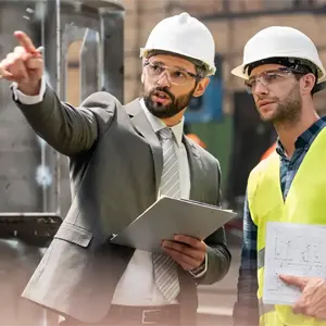 An external setting where a man wearing a suit and a hard hat is pointing something out to another man wearing a hard hat and a hi-viz jacketAn external setting where a man wearing a suit and a hard hat is pointing something out to another man wearing a hard hat and a hi-viz jacket