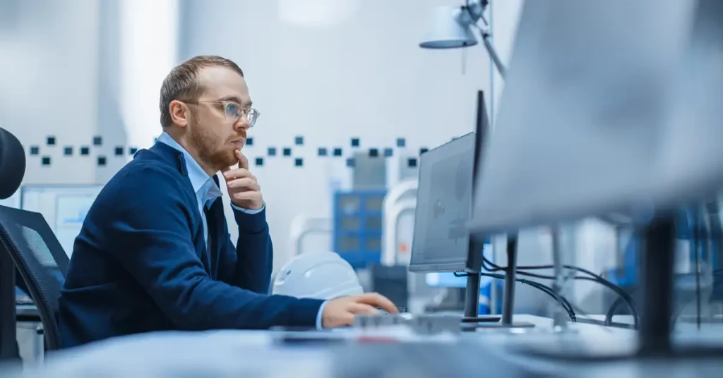 A man wearing glasses and informal work clothes sits in front of a computer