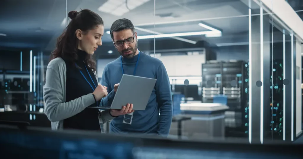 A woman and a man stand looking at a laptop help by the woman with an office IT server room in the background