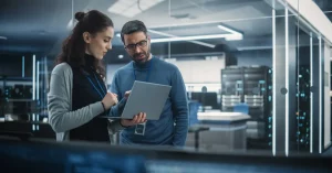A woman and a man stand looking at a laptop help by the woman with an office IT server room in the background