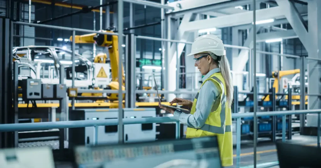 A woman in a white hard hat and yellow hi-viz jacket checks something on a laptop standing in an industrial environment