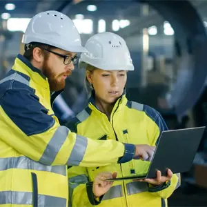 A man and a woman in hard hats and hi-viz jackets discuss something on a laptop in a factory environment