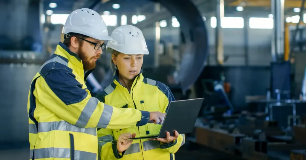 A man and a woman in hard hats and hi-viz jackets discuss something on a laptop in a factory environment