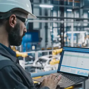 A man wearing a hard hat works on a laptop in an industrial environment