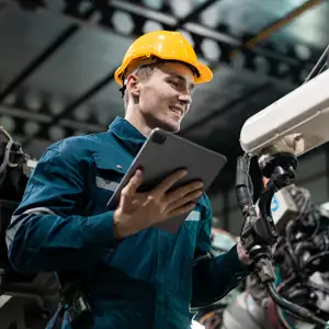 A man in a yellow hard hat holds a tablet in one hand whilst examining a piece of equipment in a factory